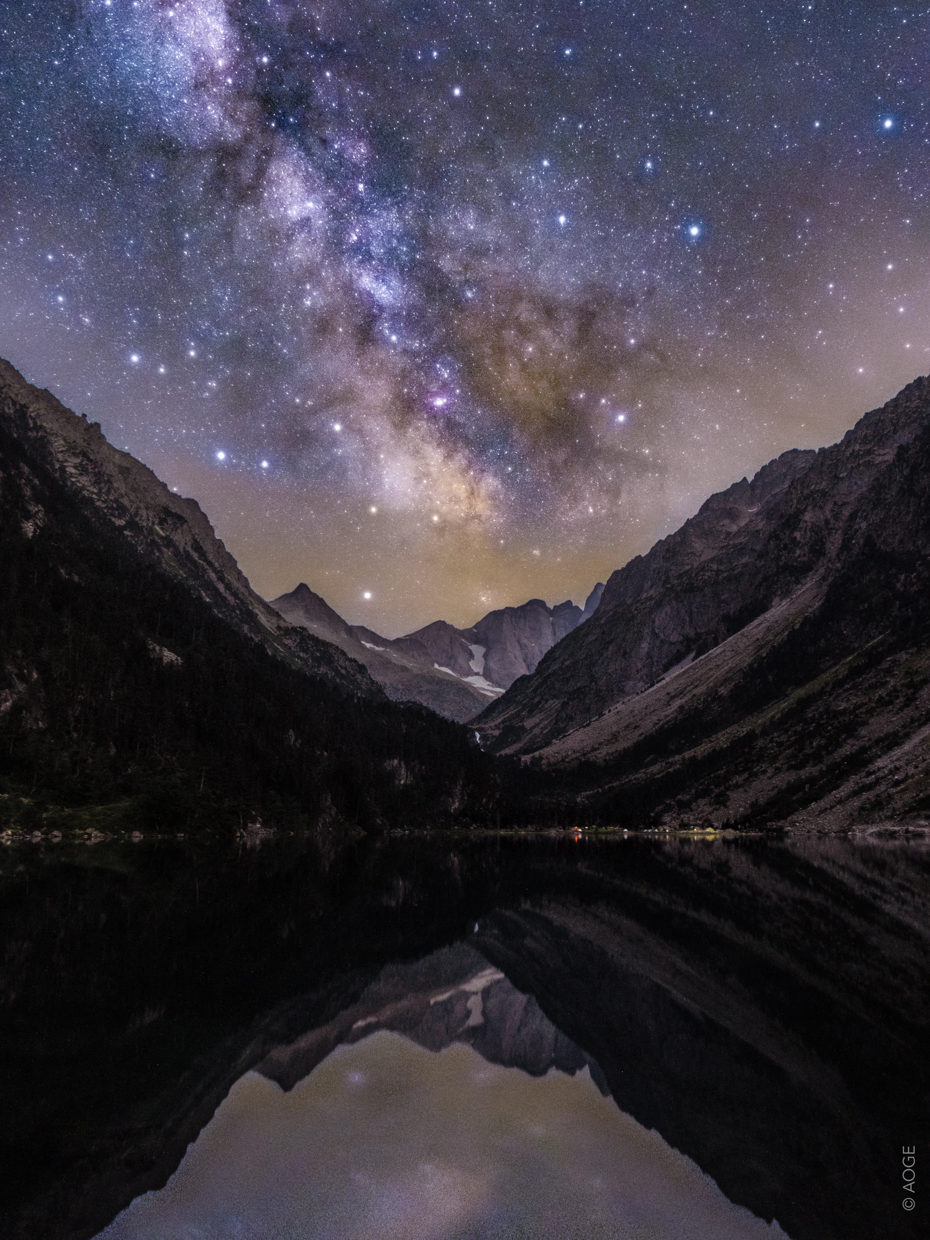 The Milky Way arching over a mountain valley, with stars reflected in a high-altitude lake