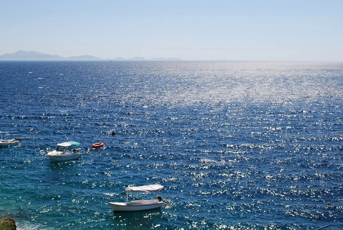 Small boats moored on calm, deep-blue Adriatic Sea water with distant islands low on the hazy horizon under a clear sky