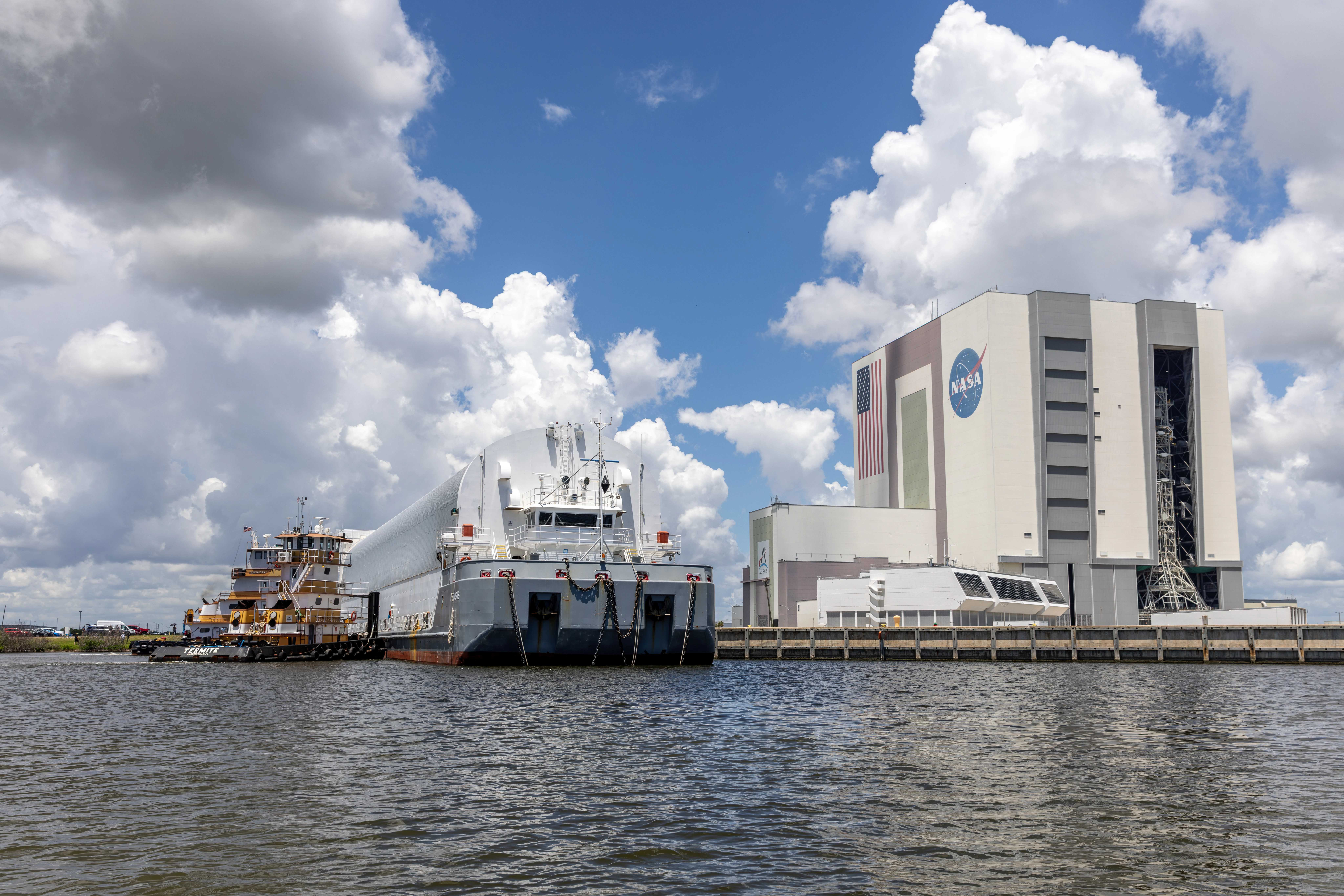 NASA's Pegasus barge, carrying the Roman Space Telescope in a white shipping container, moored at the Launch Complex 39 turn basin with the Vehicle Assembly Building behind it