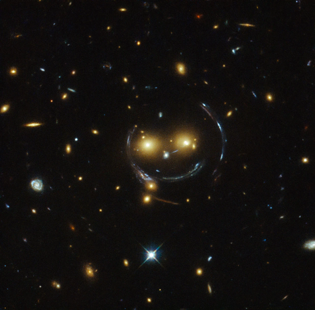 Hubble image of galaxy cluster SDSS J1038+4849: two bright yellow galaxies flanked by curved blue arcs of strong gravitational lensing that form a smiling-face pattern, against a field of faint distant galaxies.