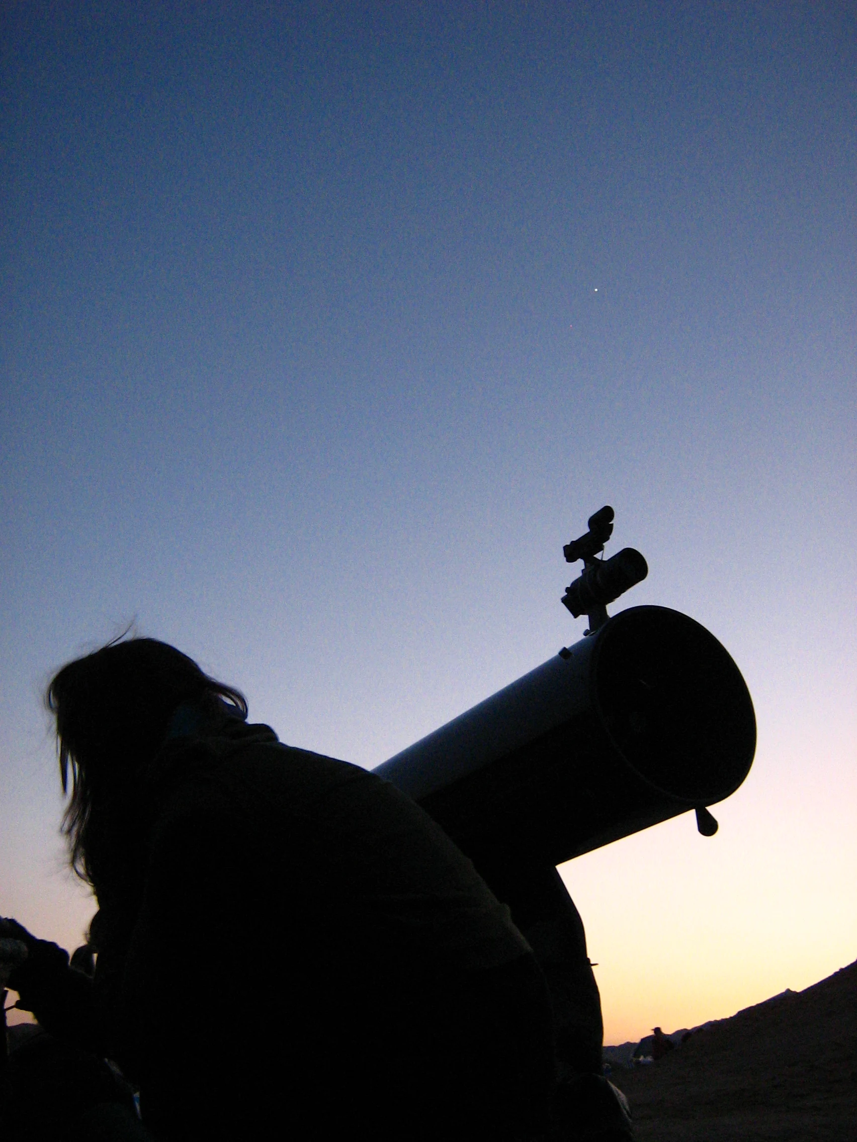 Silhouette of an amateur astronomer beside a Newtonian reflector telescope against deep-blue desert twilight.