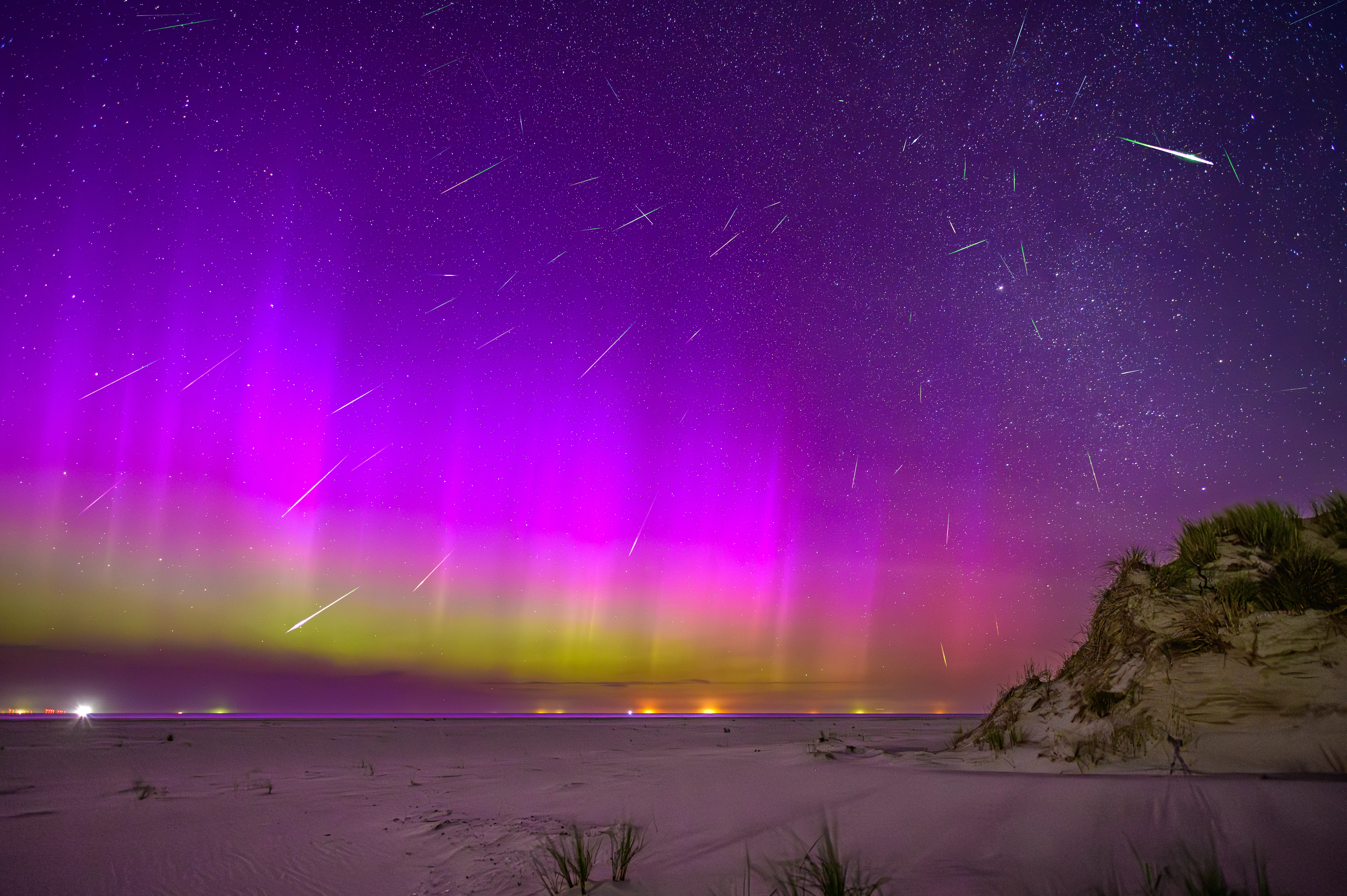 A composite of dozens of Perseid meteors radiating across the night sky over the beach at Norderney, Germany, with green aurora glowing along the horizon