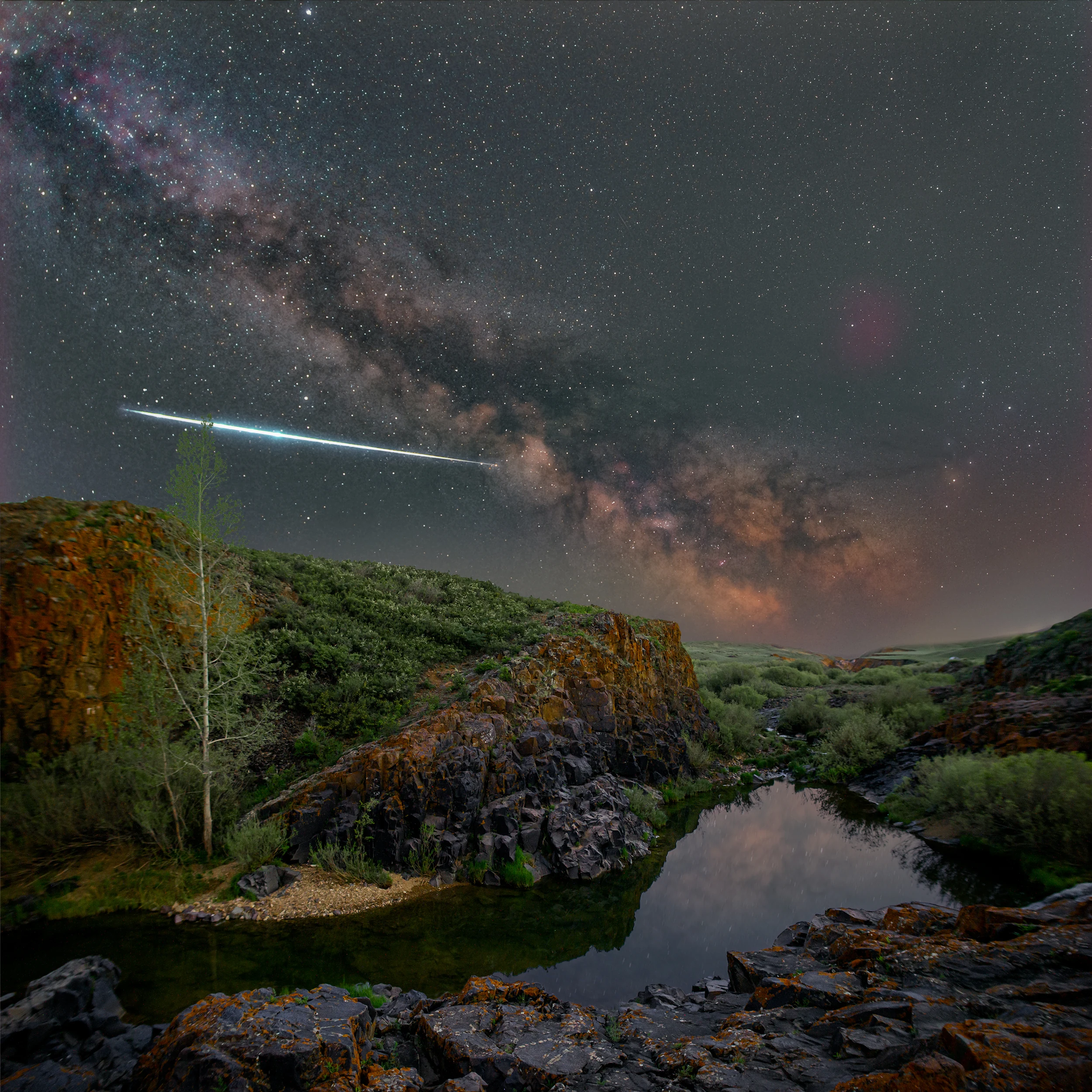 A bright bolide with a blue-green trail streaks across the Milky Way over a rocky canyon and a still reflecting pool.