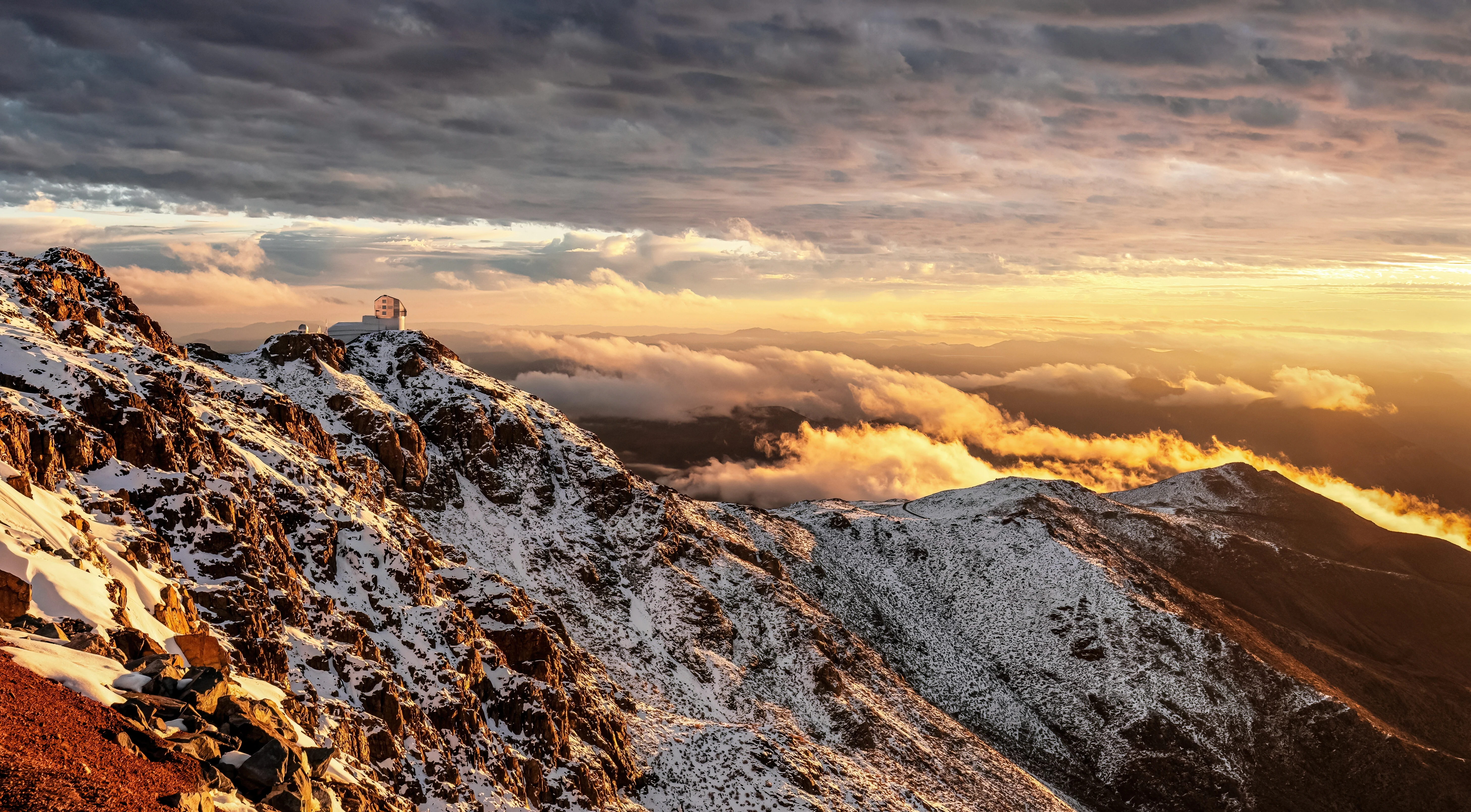 The Vera C. Rubin Observatory on a snow-covered ridge of Cerro Pachón, Chile, above a sea of clouds at sunset