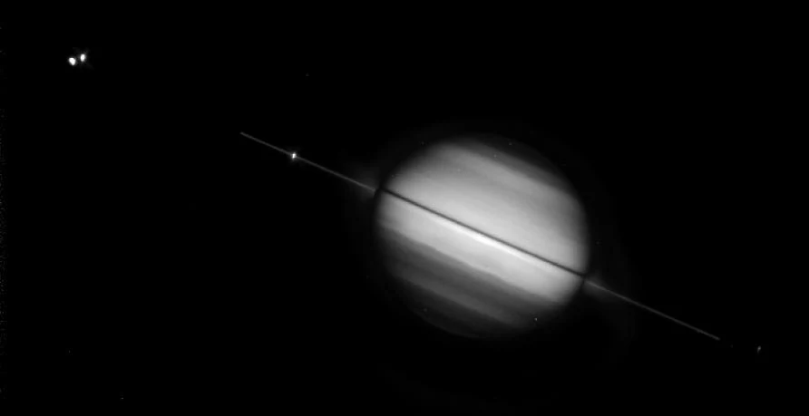 Saturn imaged by the Hubble Space Telescope with its rings turned nearly edge-on, appearing as a thin bright line across the planet's disc, with two of its moons as bright points at upper left.