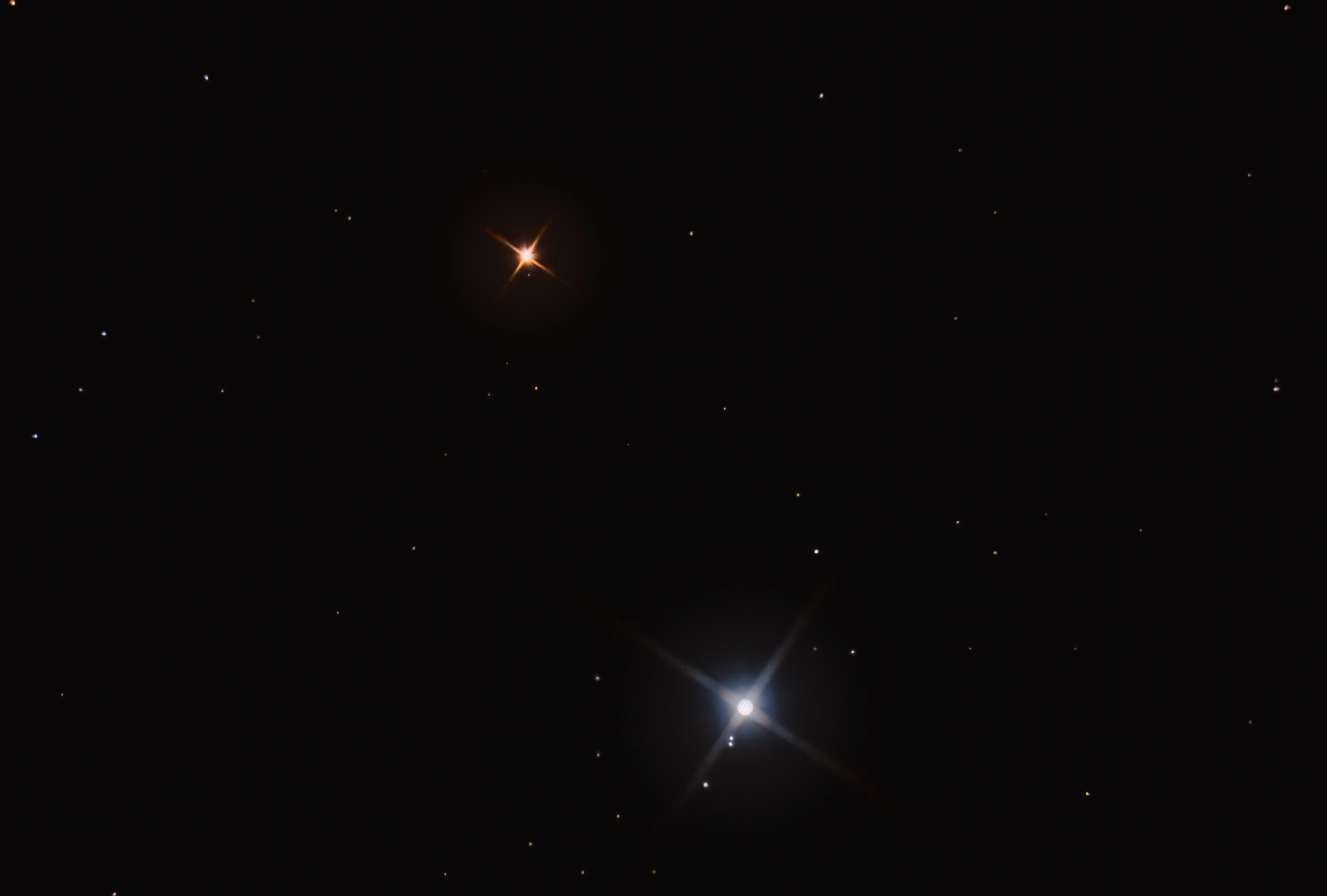 Telescope photograph of Mars and Jupiter close together on a black star field: Mars is a small orange point near the top, Jupiter a brighter blue-white point lower down with tiny dots of its moons beside it, both with four-pointed diffraction spikes.