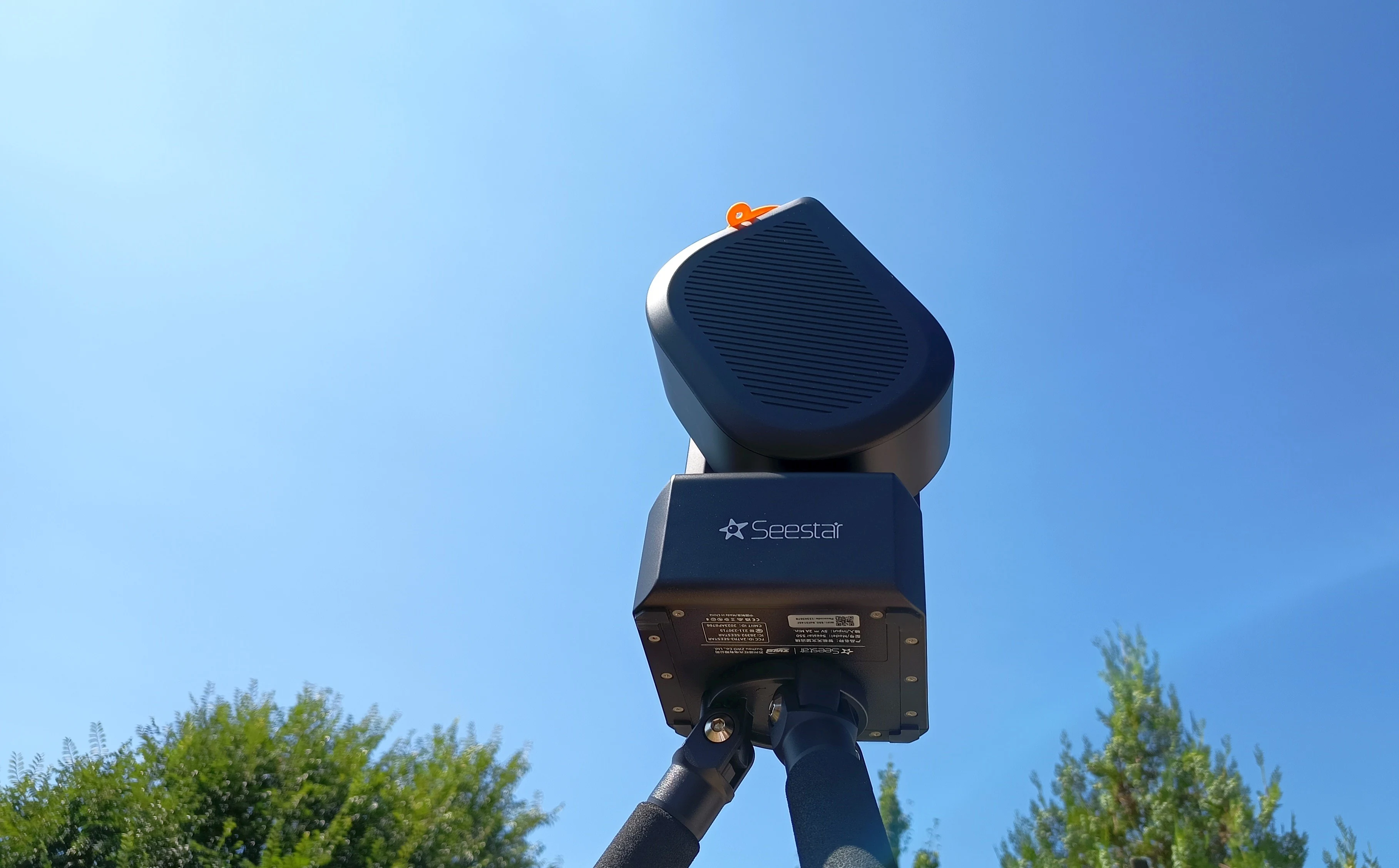 The original ZWO Seestar S50 smart telescope on its tripod, photographed from below against a clear blue daytime sky, its rounded optical tube raised above the Seestar-branded base, with treetops along the bottom edge of the frame.