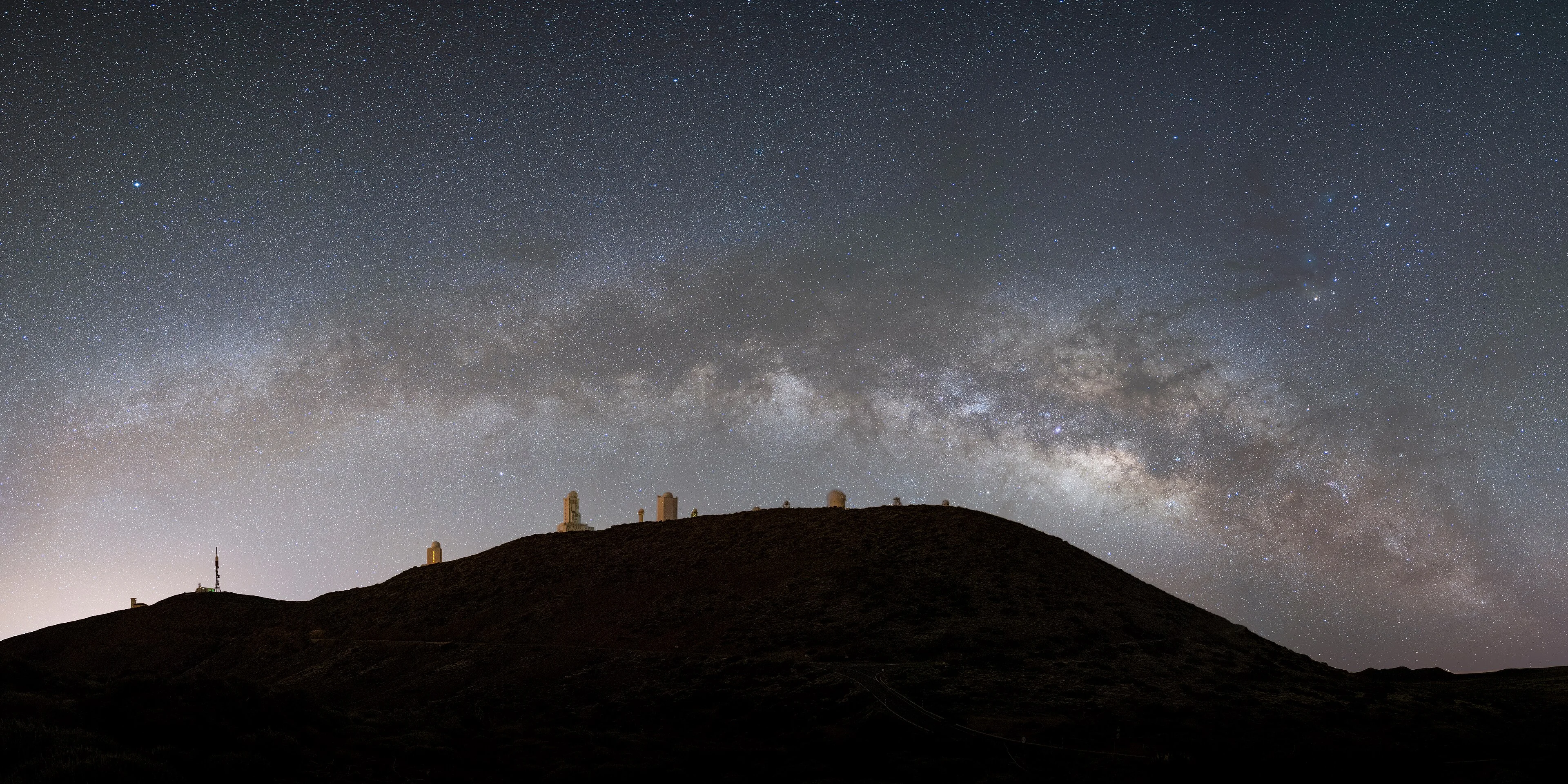 The Milky Way arching over the telescope domes of the Teide Observatory on the Izaña ridge in Tenerife, with Scorpius and Antares on the right and a faint glow of coastal light on the left horizon.
